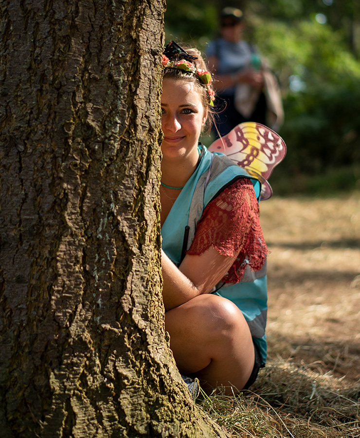 Jobs and Volunteering at Latitude Festival with Hotbox Events - Pixie volunteer smiling whilst hiding behind a tree v2022001 740x902Px72Dpi